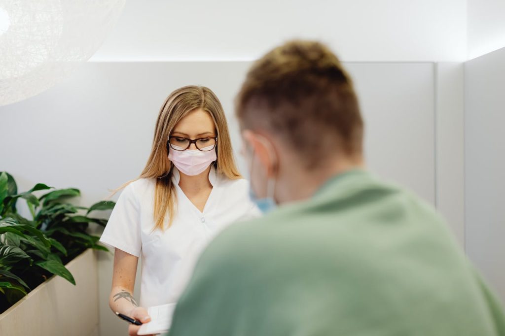 a-woman-wearing-pink-face-mask-6627859 A medical professional with a face mask talking to a patient in a clinic setting.